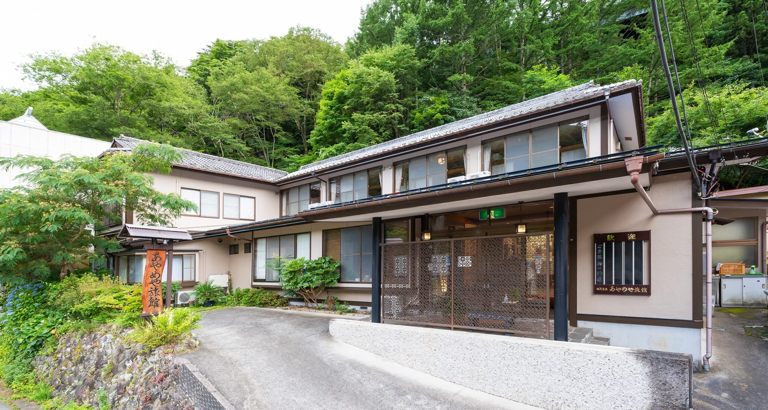 あやめや旅館 — 四万温泉・新湯地区 · 新緑の山を背にした和風の外観と石畳のアプローチ