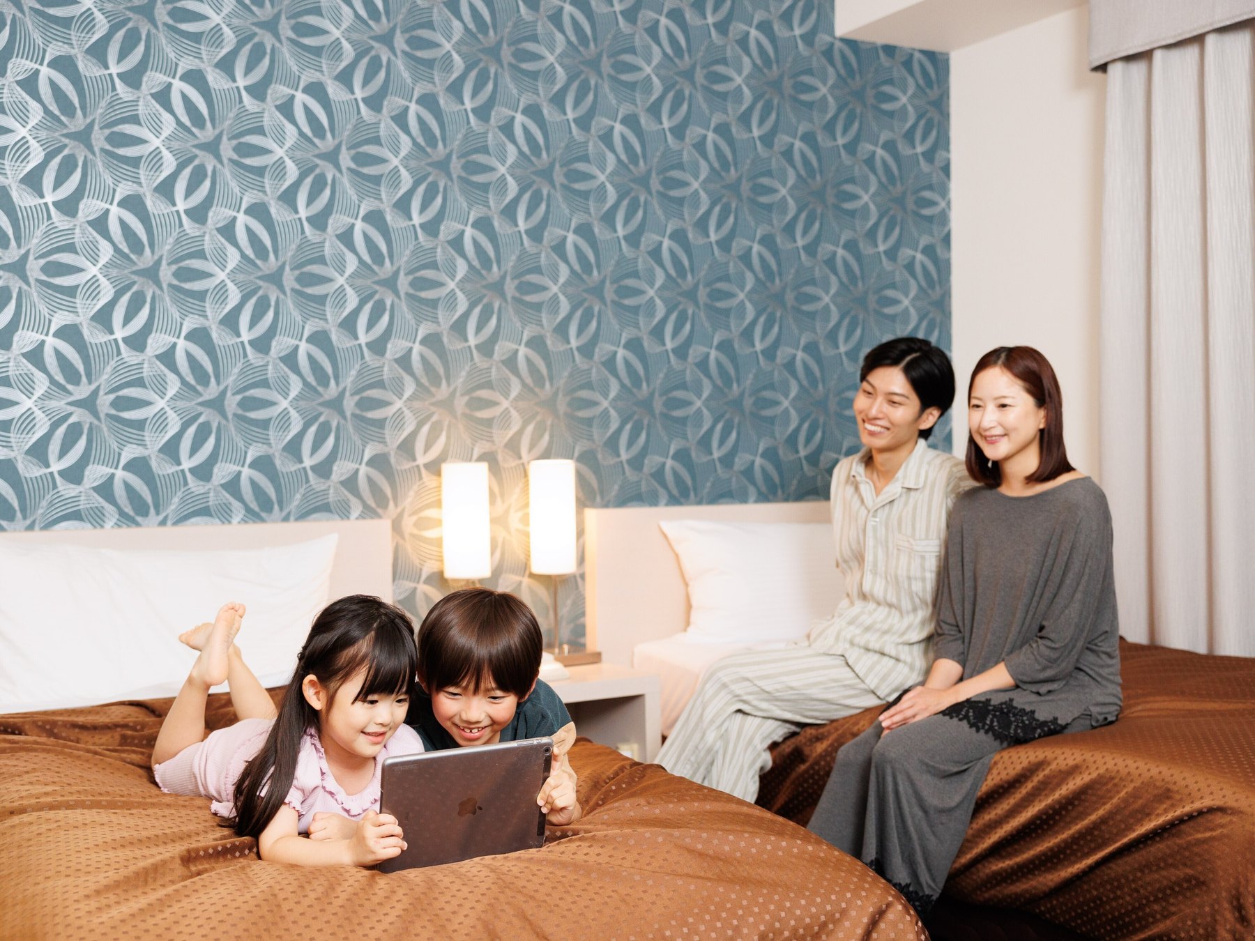 Jホテルりんくう — 常滑市・りんくう · 幾何学模様のアクセントクロスを背にした客室でくつろぐ家族