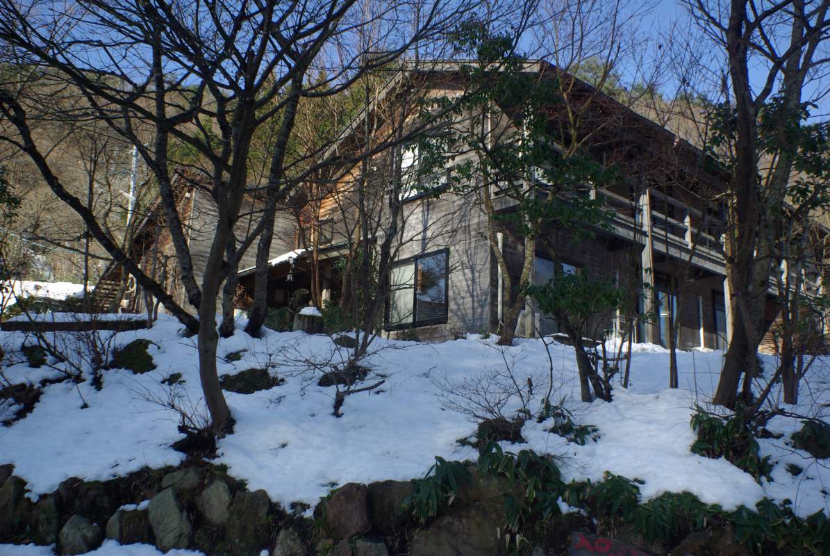 民宿たなべ — 冬の日中、山肌に残る雪と裸木に囲まれた木造宿の外観