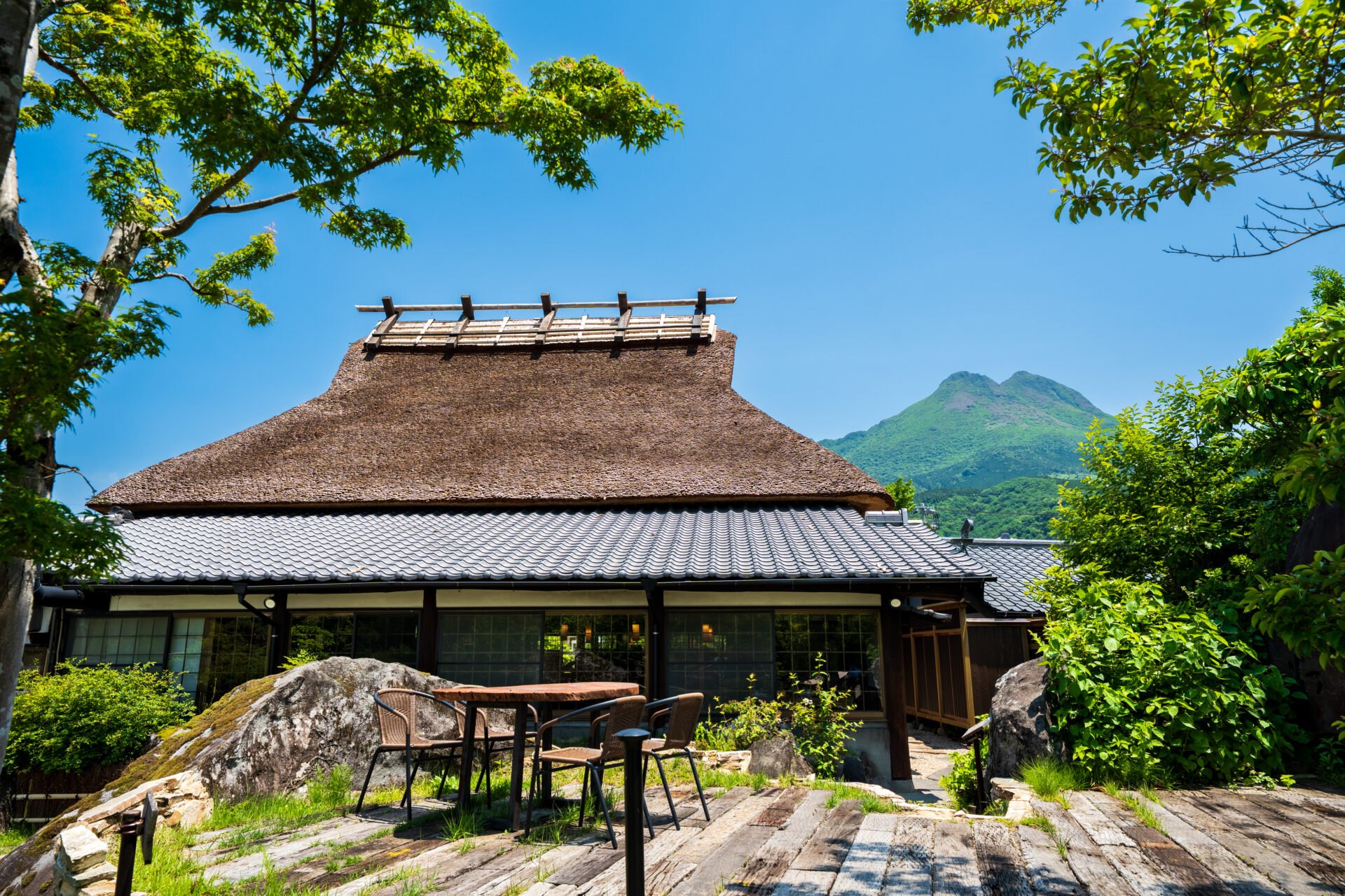 旅荘 牧場の家 — 由布市湯布院町川上 · 由布岳を背にした茅葺き屋根の母屋と庭のテラス席