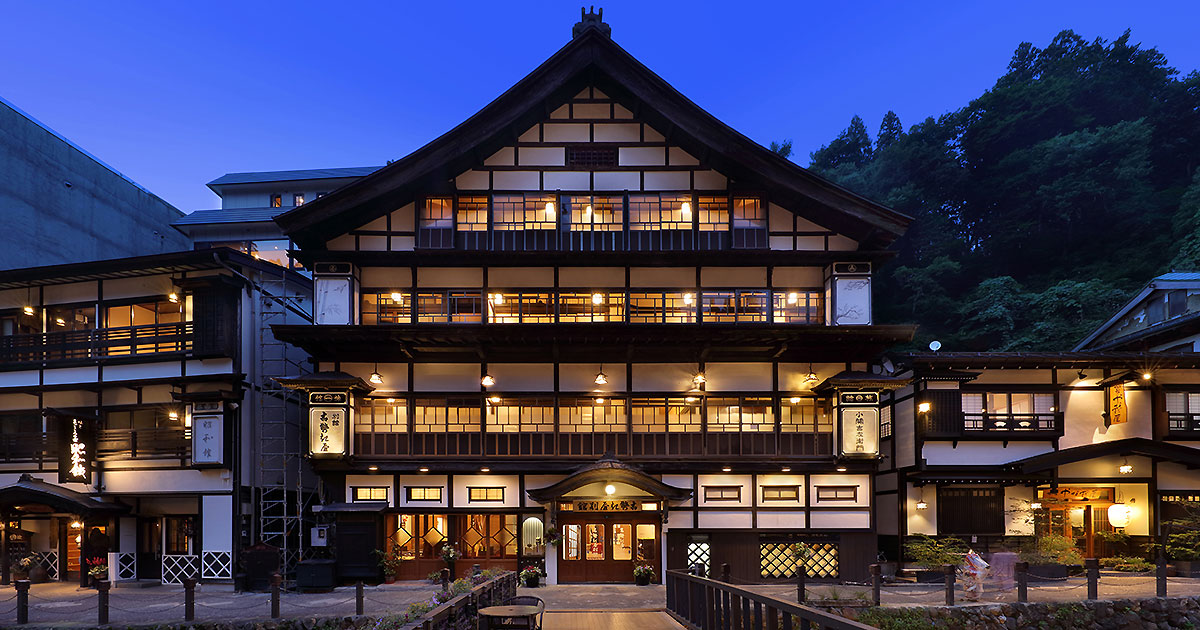 古勢起屋 別館 — 山形県尾花沢市銀山温泉 · 薄暮に灯る大正期の木造建築ファサードと銀山川沿いの遊歩道